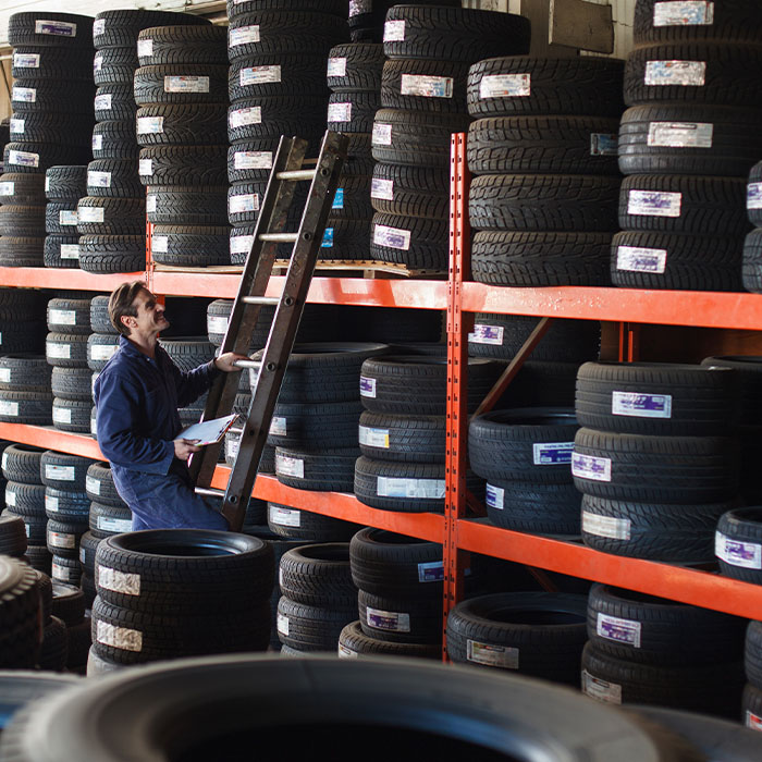 Mechanic holding a new car tire looking upward to shelf of tire inventory